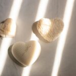 Three heart-shaped candles with textured designs basking in soft sunlight on a minimalist surface.